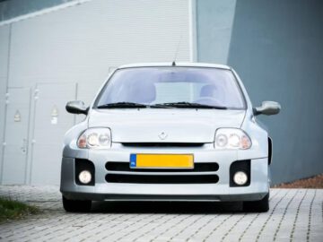 Een zilverkleurige Clio met een gele kentekenplaat, geparkeerd op een geplaveide ondergrond, met zijn gezicht naar de camera gericht. Op de achtergrond staat een grijs gebouw met deuren in industriële stijl.
