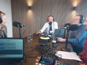 Vier personen zitten rond een tafel met microfoons en koptelefoons en zijn bezig met een AutoRAI Podcast-opnamesessie. Op de voorgrond zijn een laptop en audioapparatuur zichtbaar.