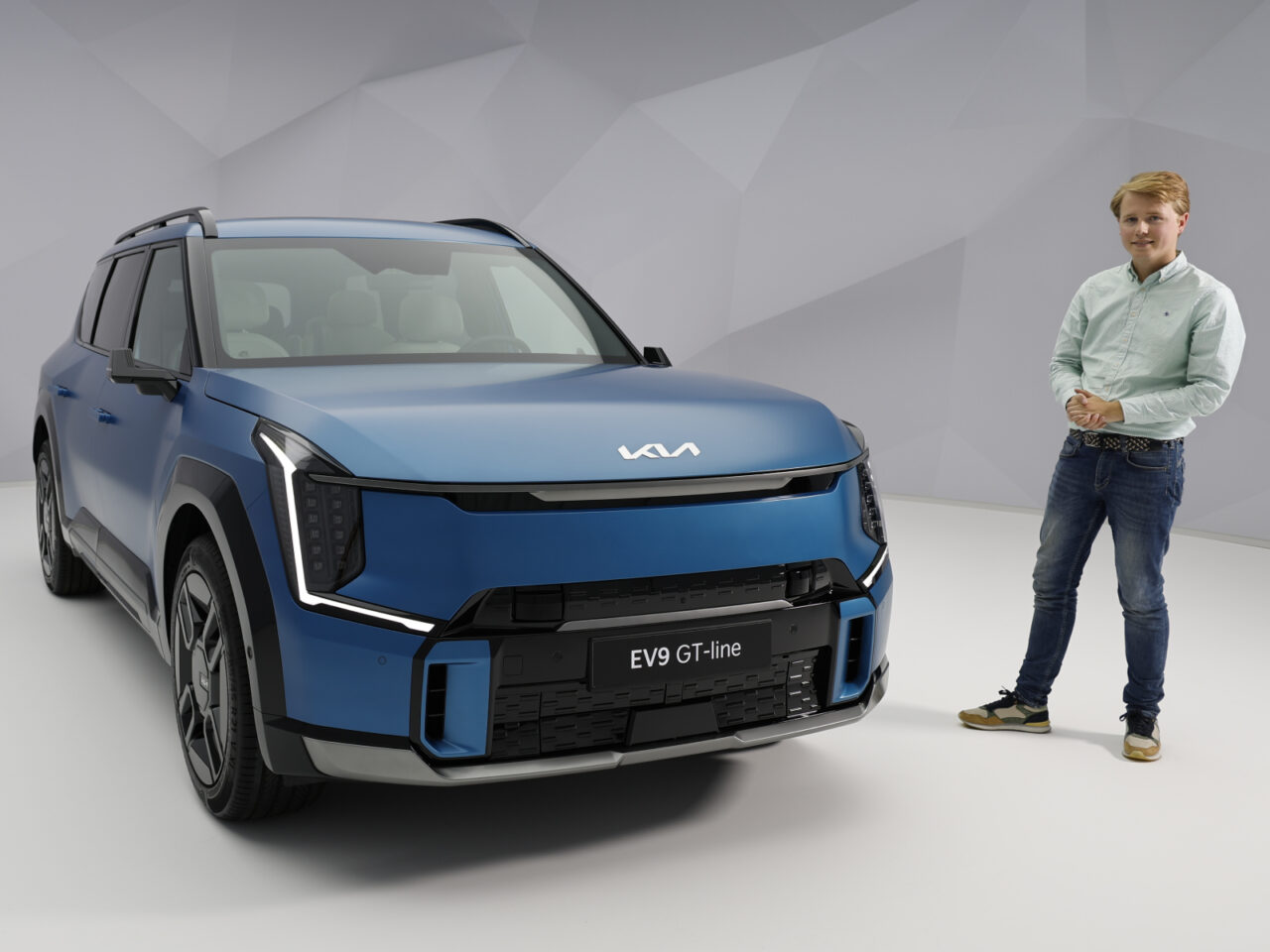 Een persoon staat naast een blauwe Kia EV9 in een studioomgeving en benadrukt het moderne ontwerp en de verfijning van de auto.