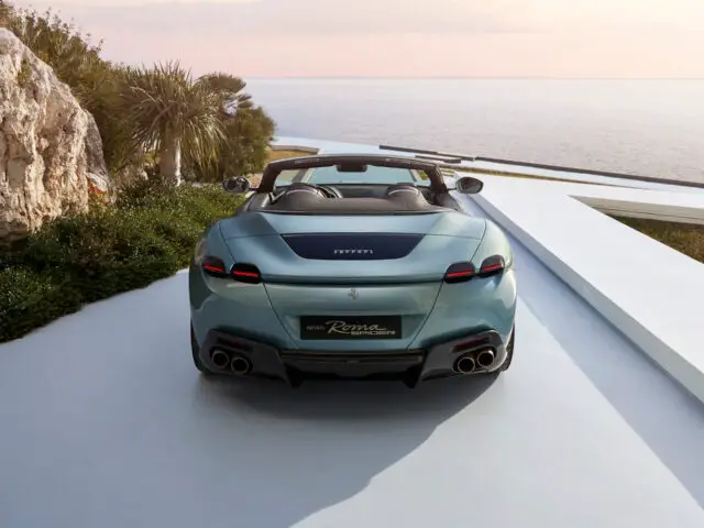 Een lichtblauwe Ferrari Roma Spider converteerbare sportwagen staat geparkeerd vlakbij een kustgezicht met palmbomen en een rotsachtige formatie op de achtergrond. De achterkant van de auto, voorzien van dubbele uitlaatpijpen en het iconische 'Ferrari'-logo, valt zichtbaar op tegen de schilderachtige achtergrond.