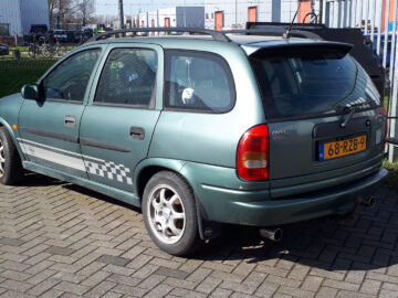 Een groene Opel Astra stationwagen met kentekenplaat "68-XZB-9" staat geparkeerd op een stenen ondergrond naast een hek en metalen gebouwen, wat doet denken aan het stoere ontwerp van de betrouwbare Opel Corsa.