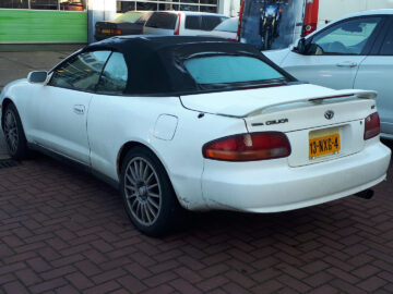 Op een drukke parkeerplaats staat een strakke witte Toyota Celica-cabriolet, met de achterkant naast andere voertuigen tentoongesteld.