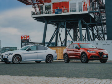 Twee auto's, een Alfa Romeo Giulia in het wit en een Stelvio in het rood, staan geparkeerd op een verhard oppervlak voor industriële machines onder een gedeeltelijk bewolkte hemel.
