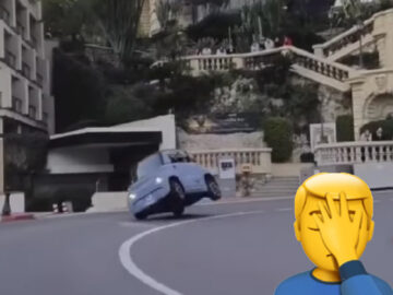 Een Citroën Ami rijdt op twee wielen terwijl hij een scherpe bocht maakt op een hellende weg nabij gebouwen en trappen, gevaarlijk wankelend voor een dreigende crash. Op de voorgrond wordt een emoji met een hand voor zijn gezicht weergegeven.