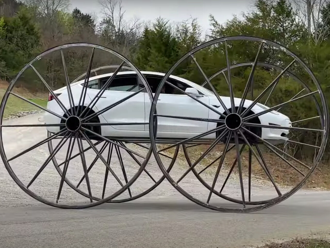 Een witte Tesla Model 3 sedan is uitgerust met extra grote dunne spaakwielen, waardoor hij een ongewoon uiterlijk krijgt. De auto staat geparkeerd op een landelijke weg met bomen op de achtergrond.