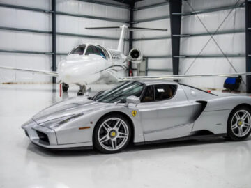 In een ruime hangar staat een Ferrari Enzo geparkeerd, met op de achtergrond een witte privéjet.