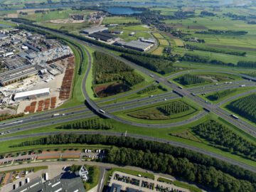 Luchtfoto van het snelwegknooppunt A27, omringd door groene velden, industriële gebouwen en schaarse woonwijken. Meerdere wegen kruisen elkaar met zichtbaar verkeer, terwijl een agrarisch landschap zich op de achtergrond uitstrekt.