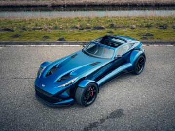 Luchtfoto van een blauwe, tweezits Donkervoort F22 sportwagen met een strak design, geparkeerd op een verharde weg naast een grasveld en waterweg.