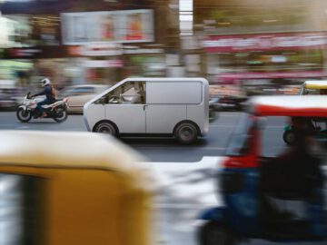 Een wit Helixx-busje rijdt door een drukke straat met een wazig bewegingseffect, geflankeerd door een motorrijder aan de linkerkant en kleurrijke tuk-tuks aan de rechterkant.