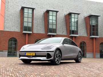 Een moderne zilverkleurige Hyundai IONIQ 6 staat geparkeerd op een bakstenen ondergrond voor een gebouw met grote ramen en rode bakstenen muren. De auto is voorzien van een elegant Europees kenteken, wat het verfijnde ontwerp benadrukt.