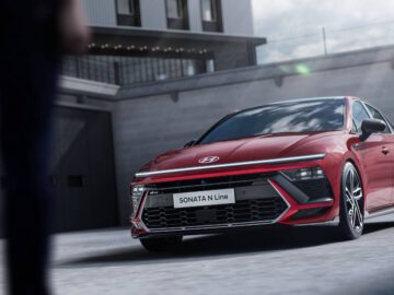 Een rode Hyundai Sonata N Line staat buiten geparkeerd voor een modern gebouw. De voorkant van de Hyundai en zijn kenmerkende grille-ontwerp worden prominent weergegeven.