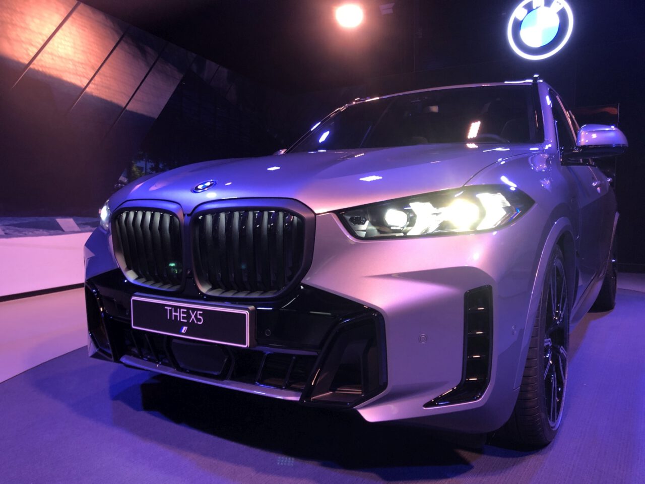 De BMW X5 SUV wordt tentoongesteld onder felle lampen en toont het vooraanzicht met verlichte koplampen en grille, in een overdekte tentoonstellingsomgeving.
