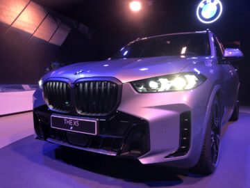 De BMW X5 SUV wordt tentoongesteld onder felle lampen en toont het vooraanzicht met verlichte koplampen en grille, in een overdekte tentoonstellingsomgeving.