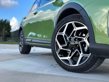 Close-up van het voorwiel van een groene Kia XCeed met een opvallende lichtmetalen velg, geparkeerd op een betonnen oppervlak onder een blauwe lucht met wolken.