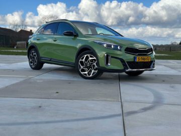 Een groene Kia XCeed SUV staat geparkeerd op een verhard terrein onder een gedeeltelijk bewolkte hemel. De auto heeft een kenteken met de tekst "S-746-FJ".