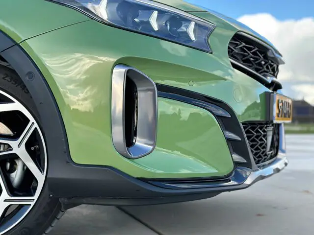 Close-up van het linkervoorgedeelte van een groene Kia XCeed, met de nadruk op de koplamp, de grille en het stuur. De foto is buiten genomen op een zonnige dag met een bewolkte hemel.