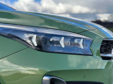 Close-up van de koplamp van een groene Kia XCeed, die het moderne, strakke ontwerp met pijlvormige LED-verlichting laat zien tegen een bewolkte hemel op de achtergrond.