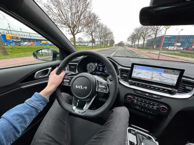 Een persoon bestuurt een Kia XCeed en houdt het stuur met één hand vast. Het dashboard en het infotainmentscherm met navigatie zijn zichtbaar. Bomen en gebouwen staan langs beide kanten van de weg verderop.