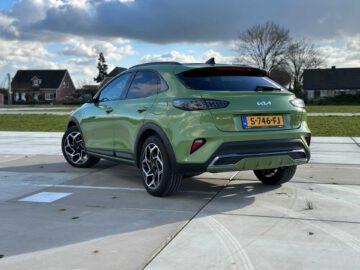 Een groene Kia XCeed compacte SUV met Nederlands kenteken staat geparkeerd op een verhard terrein in een buitenwijk, omgeven door huizen en bomen onder een gedeeltelijk bewolkte hemel.