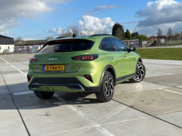 Een groene Kia XCeed crossover-SUV staat geparkeerd op een verhard terrein met een mix van bewolkte en heldere luchten op de achtergrond. De auto heeft het kenteken "S-746-FJ.