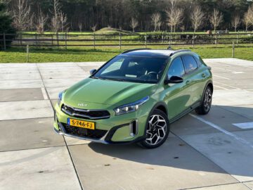 Een groene Kia XCeed SUV met schuifdak staat buiten geparkeerd op een verhard oppervlak, met een landelijk bosgebied en een omheind veld op de achtergrond.