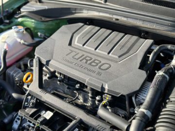 Close-up van een automotor met een deksel met het opschrift "Turbo Smartstream G" in de Kia XCeed. Rondom de kap zijn diverse motoronderdelen en slangen zichtbaar.