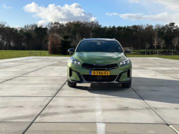 Een groene Kia XCeed met kenteken S-746-FJ staat geparkeerd op een betonnen ondergrond in een open ruimte, omlijst door bomen en een bewolkte lucht op de achtergrond.