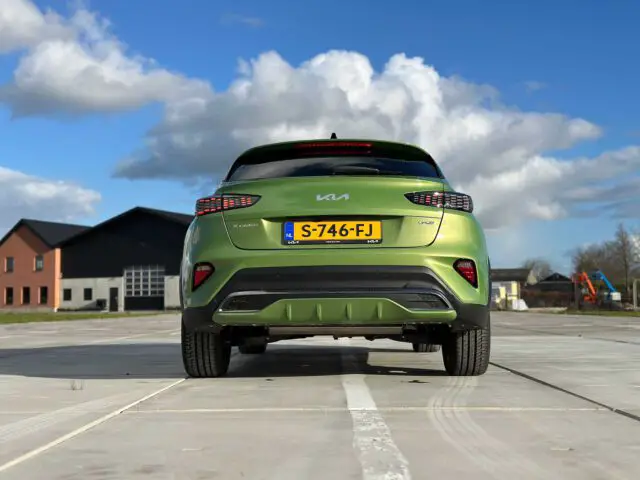 Een groene Kia XCeed met kenteken S-746-FJ staat geparkeerd op een verharde ondergrond voor twee gebouwen onder een blauwe lucht met wolken. De foto is genomen van achter de auto.