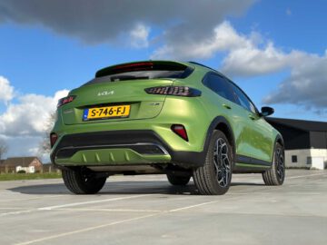 Een groene Kia XCeed met kenteken "S-746-FJ" staat geparkeerd op een betonnen ondergrond, van achteren gezien met een helderblauwe lucht en wat wolken op de achtergrond.