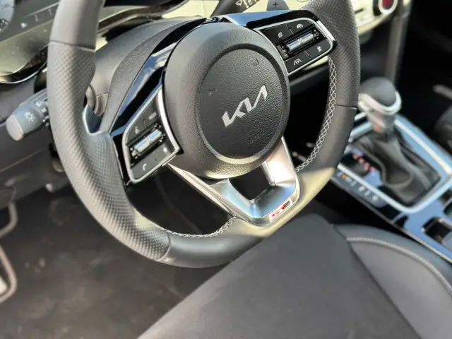 Close-up van het stuur van de Kia XCeed, met het logo, de bedieningsknoppen en een deel van het dashboard. De versnellingspook en middenconsole zijn ook gedeeltelijk zichtbaar.