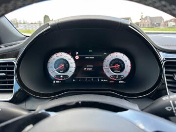 Close-up van het digitale dashboard van de Kia XCeed met een snelheidsmeter, toerenteller en verschillende indicatoren, waarbij het interieur van de auto en een buitenwijk zichtbaar zijn door de voorruit.