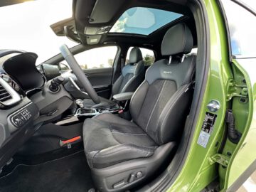 Binnenaanzicht van een Kia XCeed met zwartleren stoelen met witte stiksels, een schuifdak en een modern dashboard met verschillende bedieningselementen en displays. De deur staat open en biedt vrij zicht op de voorstoelen.