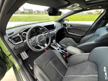 Interieur van de Kia XCeed met een strak dashboard, touchscreen-display, stuur met bedieningselementen, zwarte stoelen met witte stiksels en een schuifdak. De auto staat geparkeerd met zicht op huizen buiten.