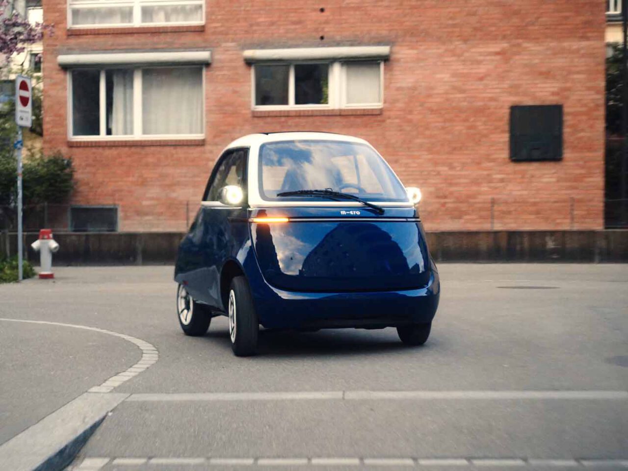 Een kleine, blauwe driewielige elektrische Microlino-auto rijdt door een stadsstraat met bakstenen gebouwen op de achtergrond.