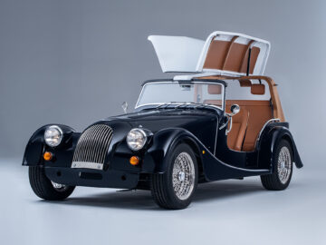 Een zwarte Morgan Plus 4 Spiaggina cabriolet in vintage-stijl met de vleugeldeuren open en een bruinleren interieur zichtbaar, tegen een effen achtergrond.