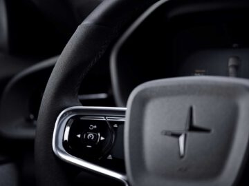 Close-up van het stuur van een auto met het Polestar-logo, bedieningsknoppen op de linker spaak en een glimp van het dashboard op de achtergrond, wat de verfijnde details van de Polestar 2 BST edition 230 benadrukt.