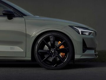 Close-up van het linker voorwiel van een Polestar 2 BST edition 230, met zwarte lichtmetalen velgen en een zichtbare oranje remklauw. De auto is lichtgrijs en heeft een strak design.