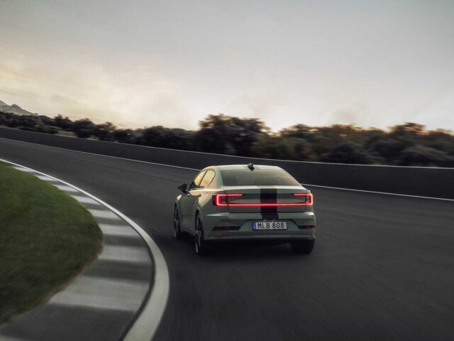 Een Polestar 2 BST edition 230 met zichtbaar kenteken "MLB 808" rijdt op een bochtige weg omzoomd door gras en bomen. De lucht lijkt bewolkt.