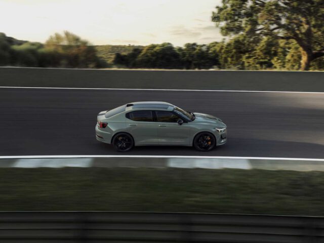 Een Polestar 2 BST edition 230 zilveren sedan rijdt op snelheid op een racecircuit, met bomen en groen op de achtergrond.