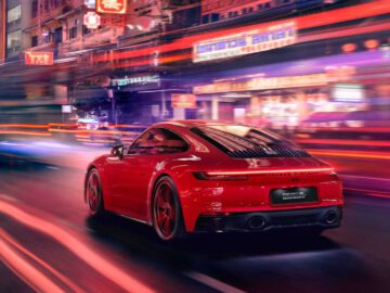 Een rode Porsche 911 Carrera GTS snelt 's nachts door een helder verlichte, drukke stadsstraat en creëert bewegingsonscherpte-effecten met de omringende lichten en borden.