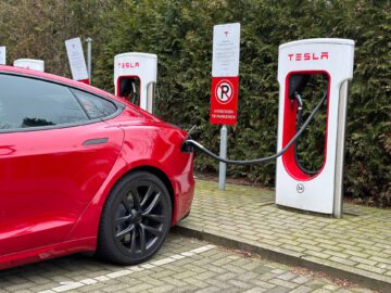 Een rode Tesla Model S Plaid laadt op bij een Tesla Supercharger-station, aangesloten op een oplader met een zwarte kabel op een door groen omgeven parkeerplaats.