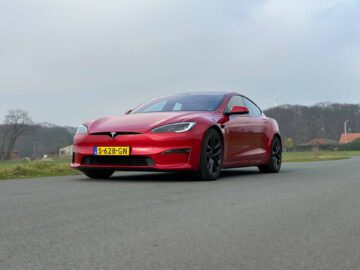 Een rode Tesla Model S Plaid staat geparkeerd op een verharde weg met een kentekenplaat zichtbaar, tegen een achtergrond van bomen en bewolkte lucht.