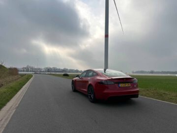 Een rode Tesla Model S Plaid staat op een bewolkte dag geparkeerd op een landelijke weg naast een veld. In de verte staat een windturbine.