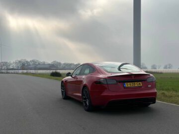 Een rode Tesla Model S Plaid geparkeerd op een landelijke weg onder een bewolkte hemel, met zonlicht dat door de wolken breekt.