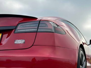 Close-up van de achterkant van een rode Tesla Model S-plaid met een zwarte spoiler, met het achterlicht, een deel van de kentekenplaat en een streepjescodesticker op de bumper.