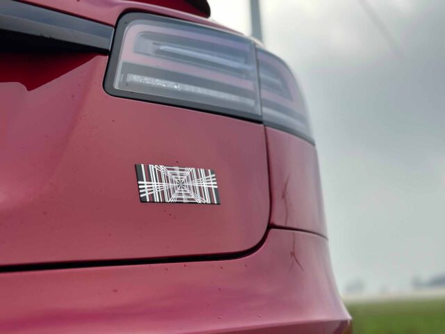 Close-up van het achterste gedeelte van een rode Tesla Model S-plaid met een geometrische zwart-witte sticker onder het achterlicht en een licht bewolkte lucht op de achtergrond.