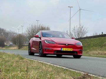 Een rode Tesla Model S Plaid rijdt op een weg met windturbines en grasvelden op de achtergrond.