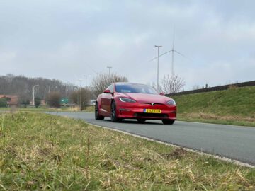 Een rode Tesla Model S Plaid met een zichtbaar kenteken rijdt op een weg, met gras en windturbines op de achtergrond onder een bewolkte hemel.