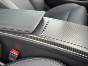 Close-up van de middenconsole van een Tesla Model S Plaid met een koolstofvezelafwerking, lederen armleuning en zwartleren stoelen. Aan de linkerkant is een rode gordelsluiting zichtbaar.