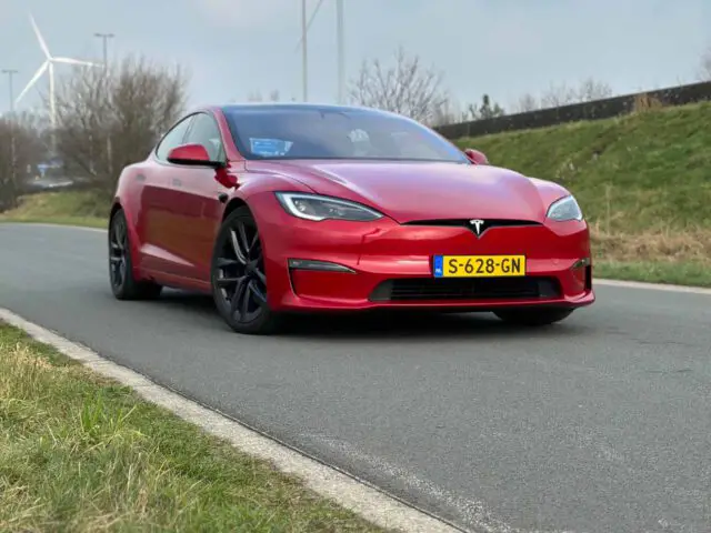 Een rode Tesla Model S Plaid met zwarte wielen staat geparkeerd op een weg. De auto heeft een Nederlands kenteken en staat buiten in de buurt van grasvelden en een windturbine.